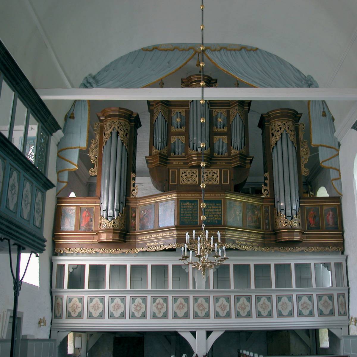 Foto der Schnitger Orgel in Steinkirchen. Die Orgel wird von einer hellblau bemalten Stuckarbeit umrahmt, die einen theaterähnlichen Vorhang darstellt. Das Blau findet sich auch im Altar, der Kanzel und den Emporen wieder..