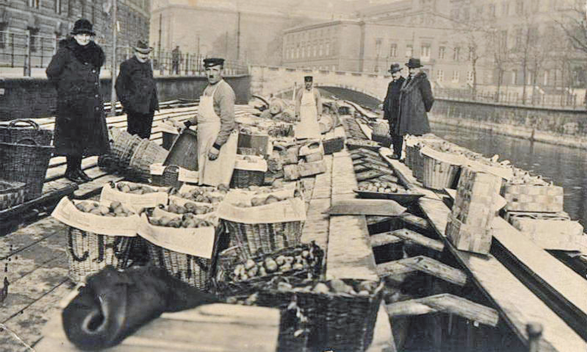 Schwarz-Weiß-Foto um 1930 eines Altländer Obsthändlers auf seinem Kahn in Berlin. Kunden betrachten das Obst, das in Siften angeboten wird.